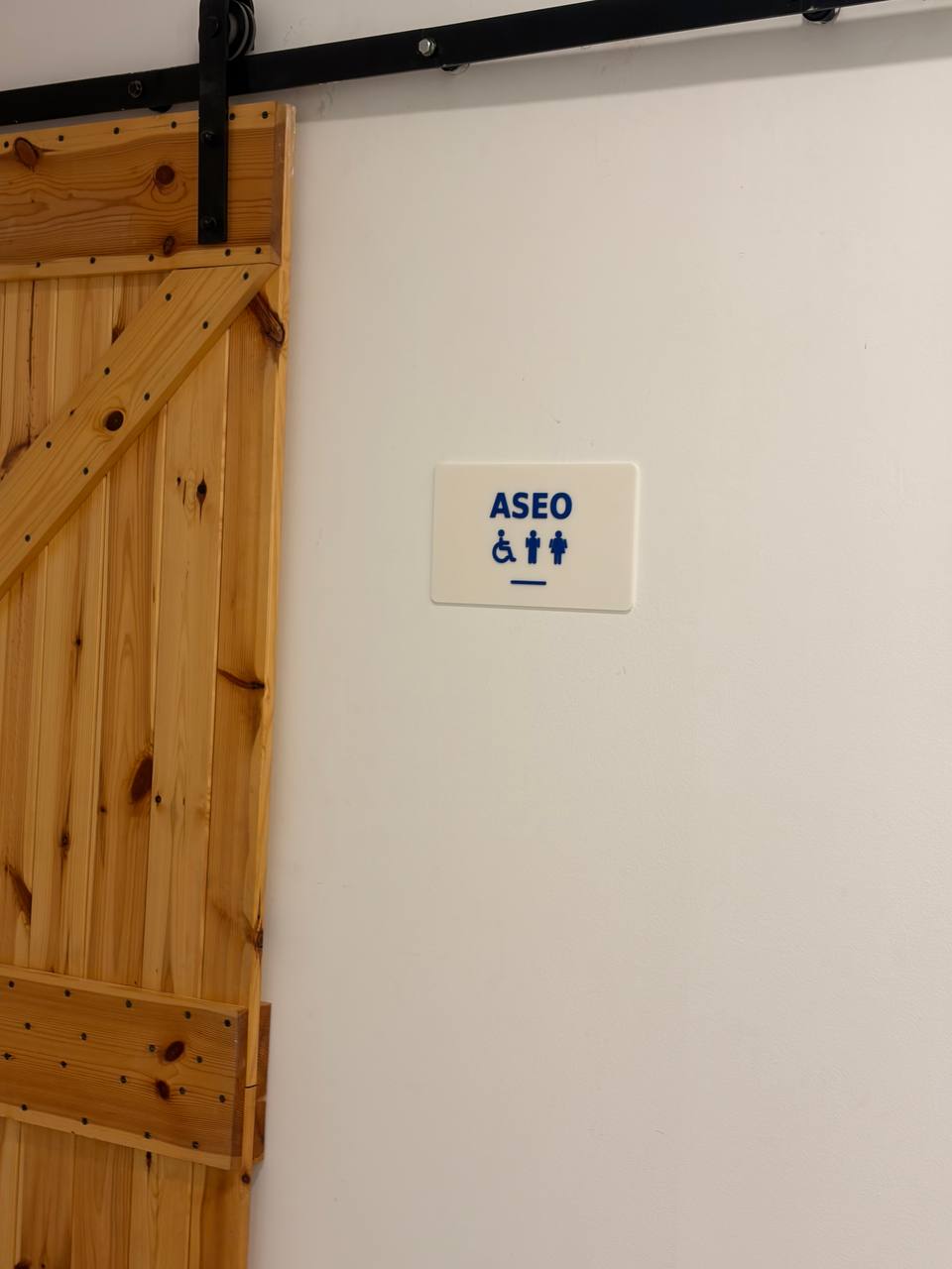 Rótulo blanco con el texto ASEO y los símbolos de baño junto a una puerta de madera